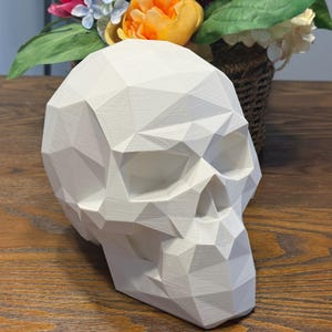 May include: A white, geometric skull sculpture with a faceted design. The skull is positioned on a wooden surface, with a floral arrangement in the background. The sculpture has a modern, minimalist aesthetic.