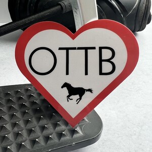 May include: A red heart-shaped sign with the text "OTTB" and a black silhouette of a horse running inside the heart.