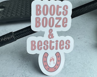 以下が含まれることがあります： ピンクの文字で「Boots Booze & Besties」と書かれた白いステッカー。ピンクの馬蹄形の中に、シャンパングラスが2つ入っています。