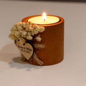 K&ouml;nnte beinhalten: Eine brennende Kerze in einem Holzhalter. Die Kerze sitzt in einer kreisf&ouml;rmigen Aussparung. Der Halter ist mit Trockenblumen und einem Holzherz mit eingraviertem Text verziert. Die Kerzenflamme ist hell.