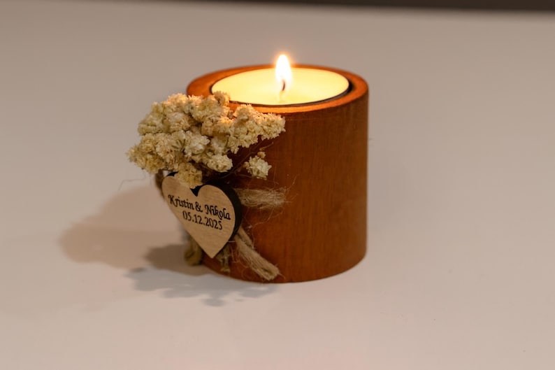 K&ouml;nnte beinhalten: Eine brennende Kerze in einem Holzhalter. Die Kerze sitzt in einer kreisf&ouml;rmigen Aussparung. Der Halter ist mit Trockenblumen und einem Holzherz mit eingraviertem Text verziert. Die Kerzenflamme ist hell.