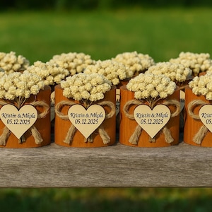 K&ouml;nnte beinhalten: Eine Sammlung kleiner, zylindrischer Holzbeh&auml;lter, die jeweils mit einem Strau&szlig; getrockneter Blumen verziert sind. An jedem Beh&auml;lter ist ein Holzherz mit dem Text "Kristin & Mkola 05.12.2025" mit Bindfaden befestigt. Die Beh&auml;lter sind auf einer Holzoberfl&auml;che angeordnet.