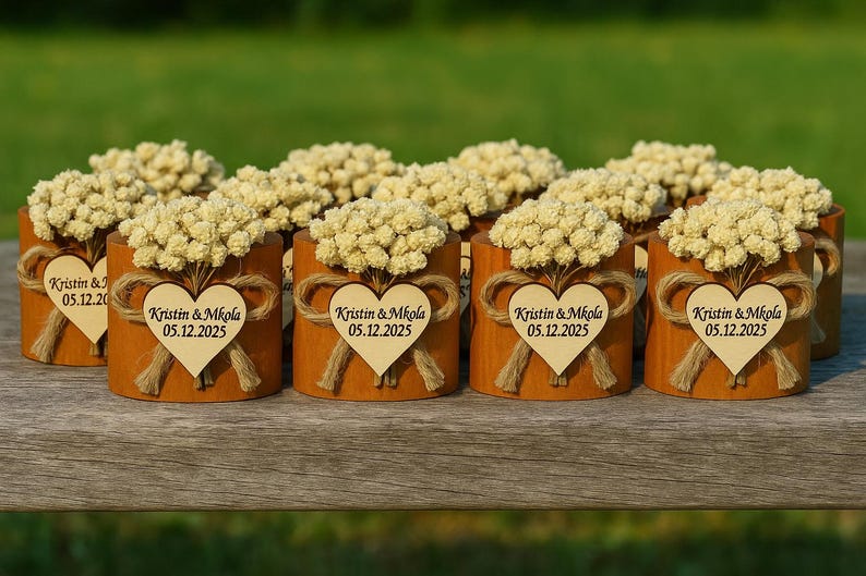 K&ouml;nnte beinhalten: Eine Sammlung kleiner, zylindrischer Holzbeh&auml;lter, die jeweils mit einem Strau&szlig; getrockneter Blumen verziert sind. An jedem Beh&auml;lter ist ein Holzherz mit dem Text "Kristin & Mkola 05.12.2025" mit Bindfaden befestigt. Die Beh&auml;lter sind auf einer Holzoberfl&auml;che angeordnet.