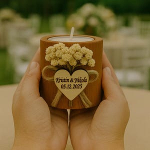 K&ouml;nnte beinhalten: Ein zylindrischer Kerzenhalter aus Holz, verziert mit einem kleinen Strau&szlig; getrockneter Blumen und einem herzf&ouml;rmigen Holzanh&auml;nger. Der Anh&auml;nger ist mit den Namen "Kristin & Nikola" und dem Datum "05.12.2025" graviert. Eine brennende Kerze befindet sich im Inneren.