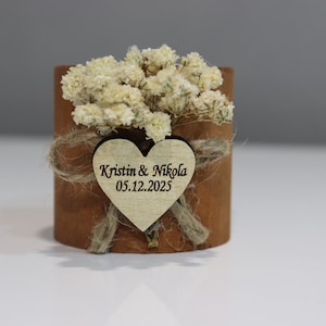 K&ouml;nnte beinhalten: Kleines, zylindrisches Holzgeschenk mit herzf&ouml;rmigem Anh&auml;nger. Der Anh&auml;nger ist mit den Namen "Kristin & Nikola" und dem Datum "05.12.2025" graviert. Das Geschenk ist mit Trockenblumen und Bindfaden verziert, passend f&uuml;r eine Hochzeit oder ein besonderes Ereignis.