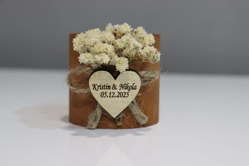 K&ouml;nnte beinhalten: Kleines, zylindrisches Holzgeschenk mit herzf&ouml;rmigem Anh&auml;nger. Der Anh&auml;nger ist mit den Namen "Kristin & Nikola" und dem Datum "05.12.2025" graviert. Das Geschenk ist mit Trockenblumen und Bindfaden verziert, passend f&uuml;r eine Hochzeit oder ein besonderes Ereignis.