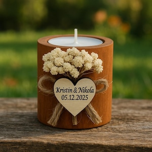 K&ouml;nnte beinhalten: Ein h&ouml;lzerner Kerzenhalter mit einer wei&szlig;en Kerze und einem kleinen Strau&szlig; wei&szlig;er Blumen. Ein Holzherz mit den Namen "Kristin & Nikola" und dem Datum "05.12.2025" ist mit Bindfaden befestigt. Der Kerzenhalter steht auf einer Holzoberfl&auml;che.