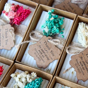 May include: Small white soap bars in brown boxes with a decorative dried flower bouquet and a thank you tag that reads "Thank You for celebrating with us Nima & Johanna 12.14.2023".