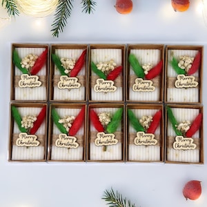 May include: Ten rectangular gift boxes with a white interior, each decorated with red and green accents, dried flowers, and a wooden "Merry Christmas" tag. The boxes are arranged on a white surface, with festive decorations.