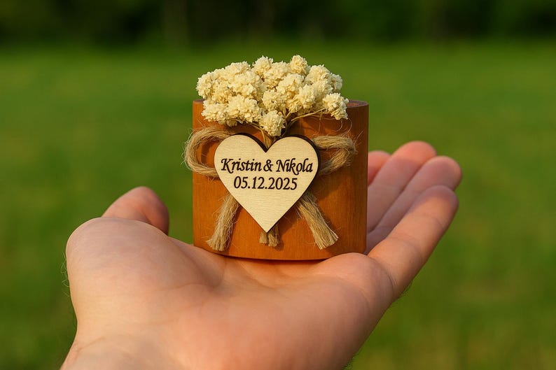 K&ouml;nnte beinhalten: Kleiner Holzbeh&auml;lter mit herzf&ouml;rmigem Anh&auml;nger und Trockenblumen. Auf dem Anh&auml;nger steht "Kristin & Nikola 05.12.2025". Der Beh&auml;lter wird in einer offenen Hand vor einem verschwommenen gr&uuml;nen Hintergrund gehalten.