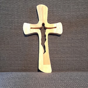 May include: A wooden cross with a unique cutout design. The cross is made of light-colored wood with a natural finish, and features a stylized figure within its form. The cross is displayed against a textured gray background.