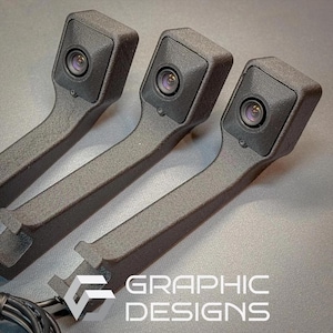 Puede incluir: Tres unidades de cámara negras y texturizadas con una lente y un brazo curvo. Las unidades están dispuestas en fila, con el texto "GRAPHIC DESIGNS" visible en la esquina inferior derecha. Un cable negro es visible en la parte inferior.