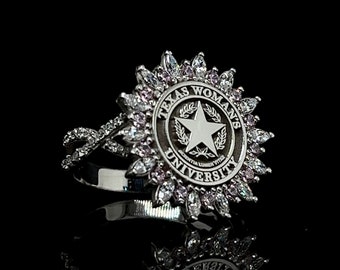 May include: A silver ring with a large, circular design featuring the Texas Woman's University logo. The logo is a star with a wreath around it, surrounded by the words "Texas Woman's University". The ring is set with small, clear and pink stones.