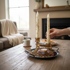 May include: A decorative arrangement on a wooden table features two tall, cream-colored candles with speckled details in ornate amber-colored candle holders. A small amber candle holder and a metal tray with decorative elements complete the display. A hand is holding one of the candles.