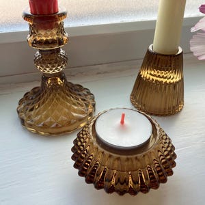 May include: A collection of amber-colored glass candle holders and a tea light holder. The candle holders have textured and patterned designs. One tall holder has a red candle, another has a cream-colored candle, and the tea light holder has a white candle.