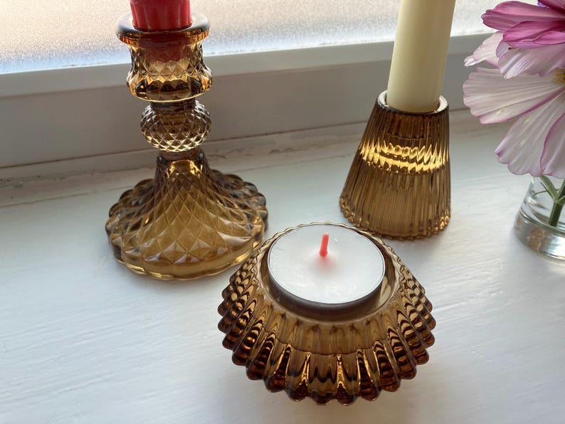 May include: A collection of amber-colored glass candle holders and a tea light holder. The candle holders have textured and patterned designs. One tall holder has a red candle, another has a cream-colored candle, and the tea light holder has a white candle.