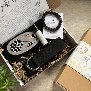 Puede incluir: Una caja de regalo de autocuidado con una zapatilla estampada en blanco y negro, una pulsera de cuentas de lava negra, aceite de aromaterapia y una correa de yoga negra. La caja incluye una tarjeta con texto sobre el autocuidado.