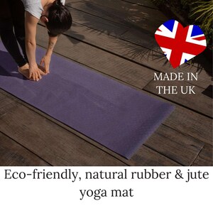 May include: A purple yoga mat made from natural rubber and jute. The mat is laid out on a wooden floor. A person is doing a forward fold yoga pose on the mat. The text "Made in the UK" is visible in the image.