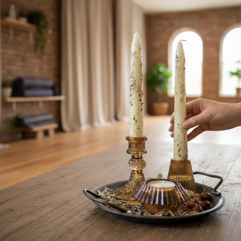 May include: Two cream-colored taper candles with speckled details sit in amber glass candle holders on a dark metal tray. A small geometric candle holder with a lit candle and decorative items are also on the tray. The scene is set on a wooden table.