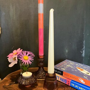 May include: A still life arrangement featuring decorative candles and floral accents. A tall, multi-colored candle stands next to a cream-colored taper candle. A small votive candle sits near a vase of flowers. Books are stacked in the background.