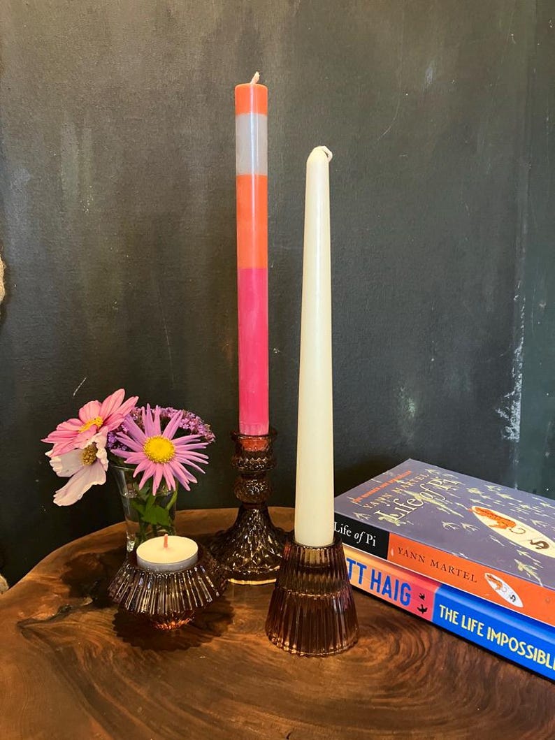 May include: A still life arrangement featuring decorative candles and floral accents. A tall, multi-colored candle stands next to a cream-colored taper candle. A small votive candle sits near a vase of flowers. Books are stacked in the background.