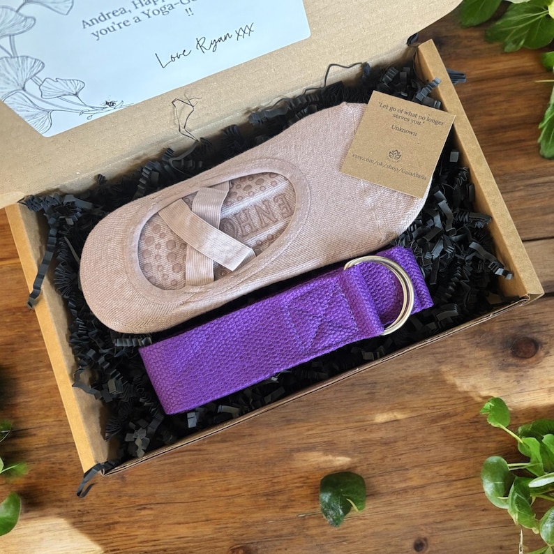 May include: A gift box filled with black shredded paper, containing a pair of beige yoga socks with a pink ribbon and a purple yoga strap with a silver buckle. The box is made of brown cardboard and has a handwritten note on the top that says "Andrea, Happy you're a Yoga! Love Ryan XXX".