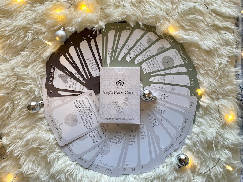 May include: A set of yoga pose cards with illustrations and instructions for beginner and intermediate poses. The cards are laid out in a fan shape on a white fluffy surface with a string of fairy lights.
