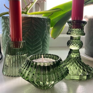 May include: Three green glass candle holders with red candles and a small candle. The candle holders are different shapes and sizes. A small candle holder is in the front, a tall one in the back, and a medium one in the middle. The background includes green plants.