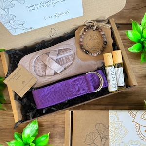 Puede incluir: Una caja de regalo llena de accesorios de yoga, que incluyen una correa de yoga morada, un par de calcetines de yoga, un brazalete marrón con una etiqueta que dice "Gaia Asana" y dos pequeñas botellas de aceites esenciales etiquetadas como "Calm" y "Energise".