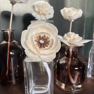 May include: A collection of reed diffuser bottles with white flower diffusers. The bottles are clear and brown, set against a dark background. The flower diffusers are a light beige colour with a brown centre.