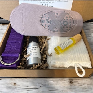 May include: A gift box filled with yoga accessories, including a purple yoga strap, a bottle of yoga mat cleaner, a white towel, a bottle of essential oil, and a pair of grey yoga socks.
