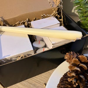 May include: A black box filled with white candles, tea lights, and packing material. A long, cream-colored candle rests on top of the other items. A small succulent plant and pine cones are in the background.