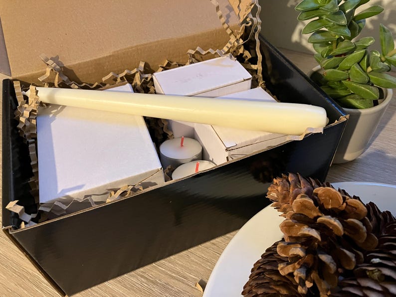 May include: A black box filled with white candles, tea lights, and packing material. A long, cream-colored candle rests on top of the other items. A small succulent plant and pine cones are in the background.