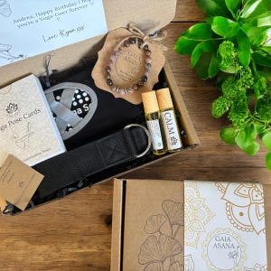 Puede incluir: Una caja de regalo con artículos de yoga. Incluye una correa de yoga, botellas de aceites esenciales, una pulsera y tarjetas de posturas de yoga. La caja también contiene una esterilla de yoga negra y una tarjeta que dice "Andrea, Happy Birthday I hope you're a Yoga-Goer forever !! Love Ryan xxx".