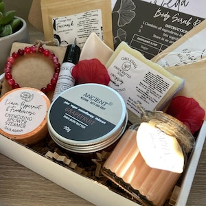 May include: A gift box filled with self-care products, including a bracelet, shea body butter, a shower steamer, soap, and a candle. The box contains a variety of items with labels and packaging, and is decorated with red accents and brown paper.