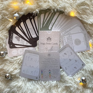 May include: A set of yoga pose cards with illustrations and instructions. The cards are laid out on a white fluffy surface with silver ornaments and string lights.