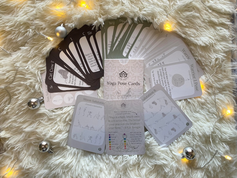 May include: A set of yoga pose cards with illustrations and instructions. The cards are laid out on a white fluffy surface with silver ornaments and string lights.