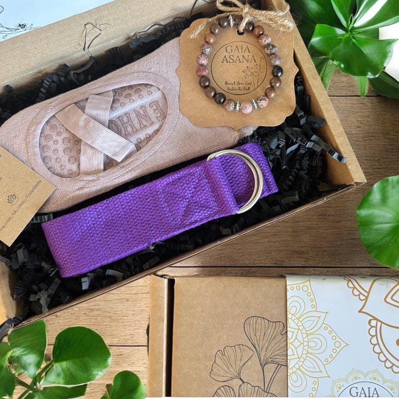 May include: A gift box filled with yoga accessories, including a purple yoga strap, a pair of beige yoga socks, and a brown gemstone bracelet with the text "GAIA ASANA" on a brown tag.