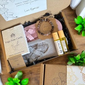 Puede incluir: Una caja de regalo llena de artículos con temática de yoga, que incluyen un juego de tarjetas de poses de yoga, un par de calcetines de yoga grises, una máscara para los ojos rosa, un brazalete con las palabras "Gaia Asana" y dos pequeñas botellas de aceites esenciales etiquetadas como "Calm" y "Energise".