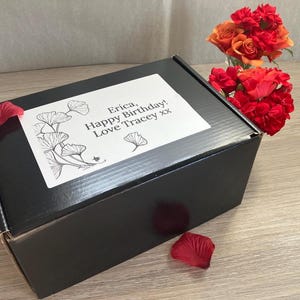 May include: A black gift box with a white label featuring a floral design and the text "Erica, Happy Birthday! Love Tracey xx." Red rose petals and a bouquet of red and orange roses are also present.