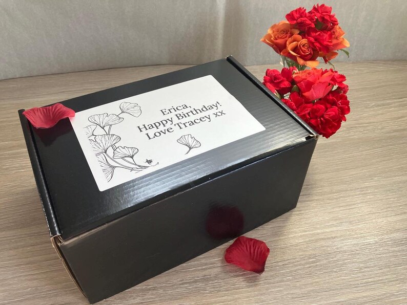 May include: A black gift box with a white label featuring a floral design and the text "Erica, Happy Birthday! Love Tracey xx." Red rose petals and a bouquet of red and orange roses are also present.