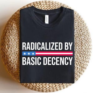 May include: Black t-shirt with the text "RADICALIZED BY BASIC DECENCY" in white block letters. A horizontal stripe with blue stars and red and white stripes is above the words "BASIC DECENCY". The shirt is displayed on a woven, circular surface.