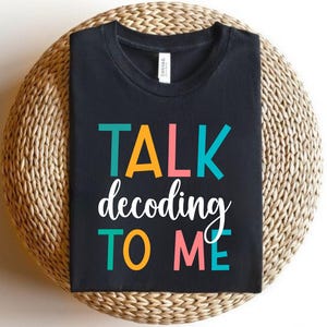Puede incluir: Camiseta negra con el texto "TALK decoding TO ME" en letras coloridas. La camiseta se muestra sobre una superficie circular tejida. El texto es en turquesa, naranja, rosa y blanco.