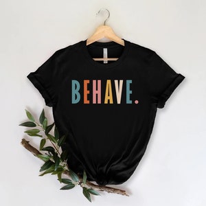 May include: Black t-shirt with the word "BEHAVE" printed in colorful letters. The letters are in shades of teal, orange, pink, yellow, and teal.