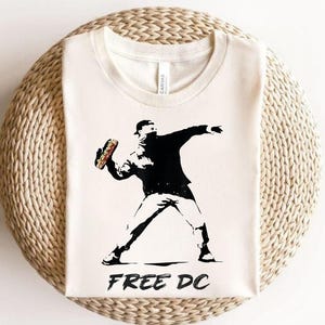 May include: Cream-colored t-shirt featuring a black and white graphic of a figure throwing an object, with the text "FREE DC" below. The image is reminiscent of street art.