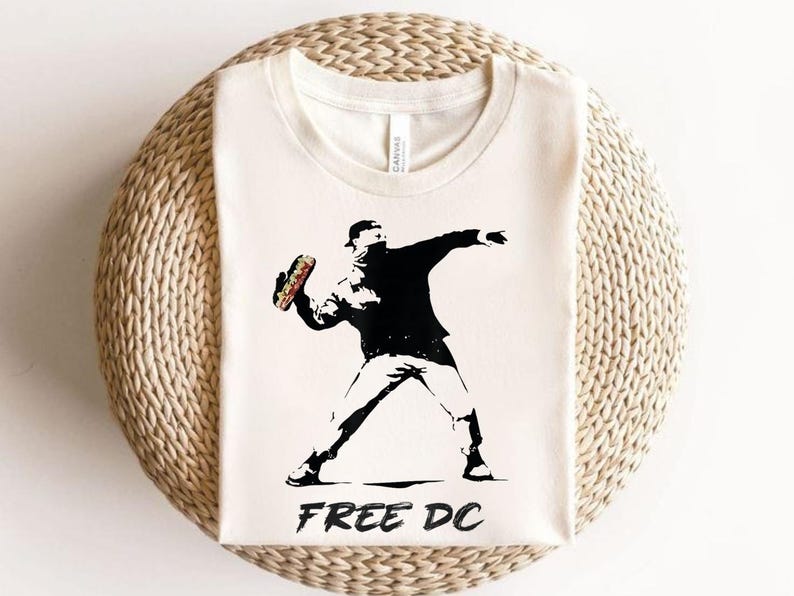 May include: Cream-colored t-shirt featuring a black and white graphic of a figure throwing an object, with the text "FREE DC" below. The image is reminiscent of street art.
