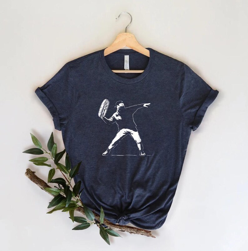 May include: Navy blue t-shirt featuring a white graphic of a person throwing a frisbee. The design is a stylized illustration. The shirt is displayed on a wooden hanger against a neutral background. The tee shirt is a casual wear item.