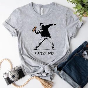 May include: A light gray t-shirt featuring a black and white graphic of a person throwing a sandwich. The text "FREE DC" is printed below the graphic. The shirt is made of a soft material.