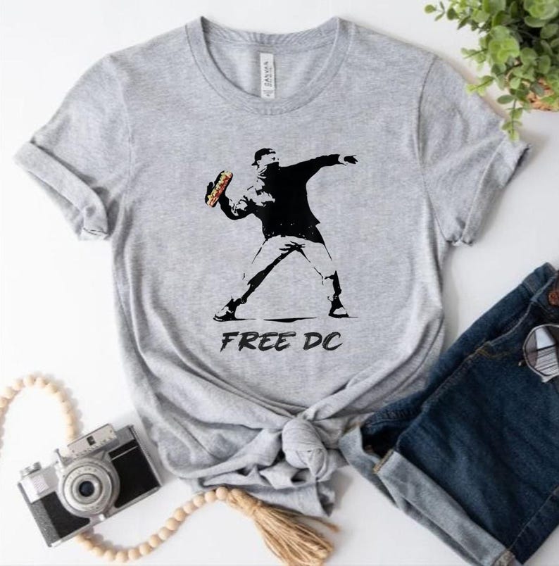 May include: A light gray t-shirt featuring a black and white graphic of a person throwing a sandwich. The text "FREE DC" is printed below the graphic. The shirt is made of a soft material.