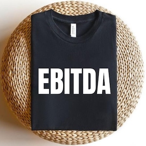 May include: Black t-shirt with the white capital letters "EBITDA" printed across the front. The shirt is displayed on a round, woven, light brown surface.